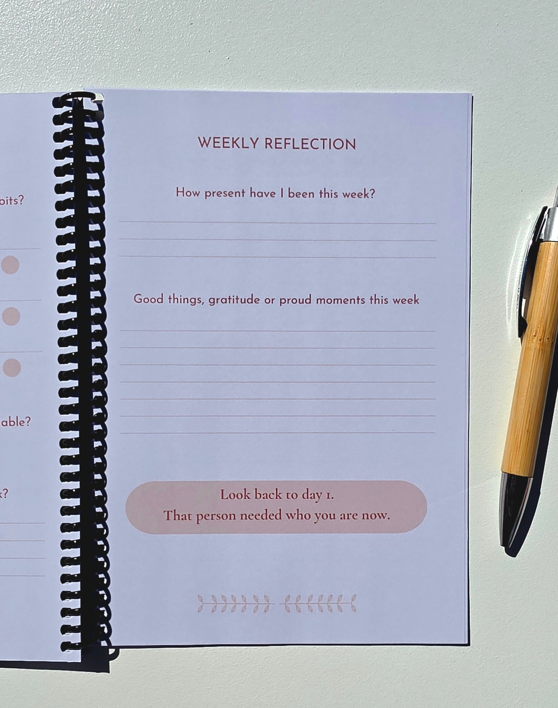 A spiral-bound weekly reflection journal page shown open on a light surface, with lined writing areas and prompts including 'WEEKLY REFLECTION' and 'Look back to day 1. That person needed who you are now.