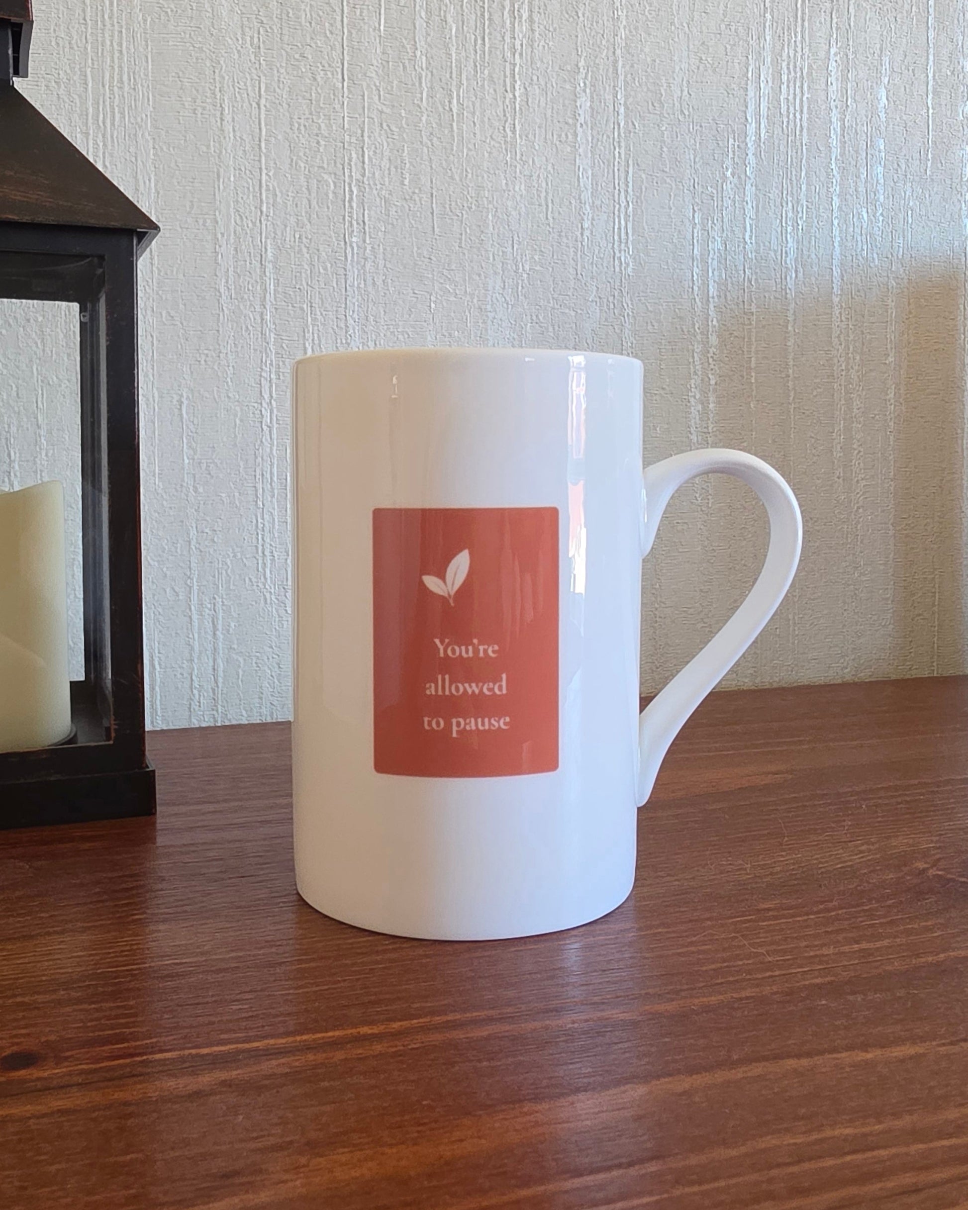 A white ceramic mug with a terracotta-colored square label on the front, sitting on a wooden table next to a dark lantern against a textured wall.